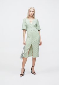 Mintgrønn kjole med pufferm, dyp V-hals, hullmønsterdetaljer og en sidespalte, kombinert med en sølvfarget håndveske og svarte hæler.