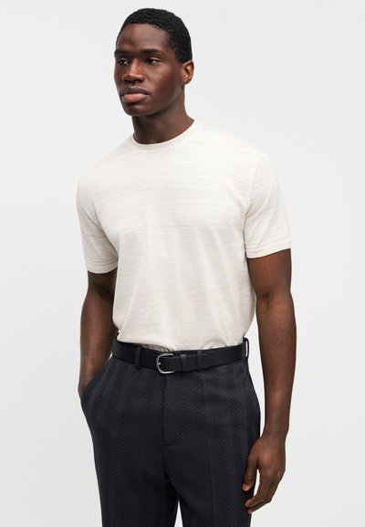 Ανοιχτό μπεζ πουκάμισο με κοντά μανίκια και διακριτική υφή, συνδυασμένο με σκούρο παντελόνι με σχέδιο και μαύρη ζώνη με ασημένια αγκράφα.