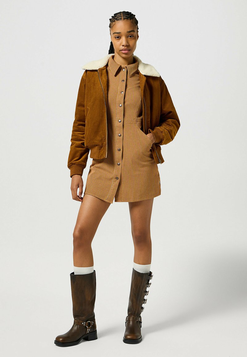 Giacca in corduroy marrone con colletto in pelliccia bianca, abbinata a un vestito camicia a scacchi. Stivali marroni al ginocchio con dettagli in argento.