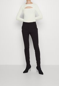 Ljus ribbad långärmad topp med en framskärning, i kombination med svarta tajta byxor och svarta klackade ankboots mot en plain bakgrund.