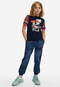 Granatowa koszulka z krótkim paskowanym rękawem z grafiką Myszki Minnie, połączona z niebieskimi dżinsami z łatą w kształcie motyla i czarnymi akcentami po bokach.