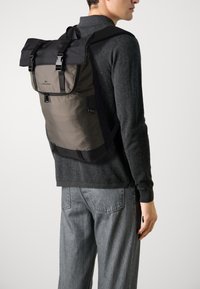 Sac à dos en tissu gris et noir, doté d'un design à enroulement, de boucles ajustables et d'une poche latérale. Texture lisse avec des matériaux contrastants.