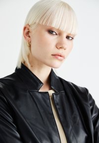 Musta nahkatakki, jossa on minimaalinen kaulusk design, sileä pinta ja vetoketjukiinnitys, päällä beige paita. Mallilla on suorat vaaleat hiukset.