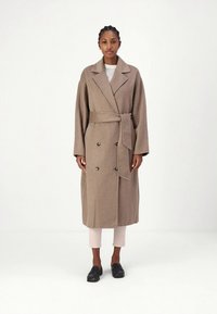 Manteau long à double boutonnage en tissu de laine beige avec une ceinture à nouer, de grands revers et des boutons à l'avant, associé à un pantalon clair et des mocassins.