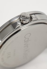 Montre en acier inoxydable argenté avec une finition polie, dotée d'un boîtier rond, d'une couronne avec le logo 'CK' et d'un fond lisse gravé.