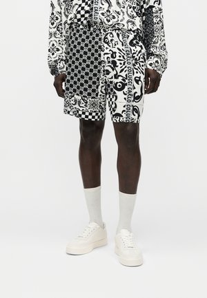 Personne portant un short à motif noir et blanc avec une chemise à manches longues assortie, des chaussettes blanches et des baskets à plateforme blanches, debout sur un fond uni.