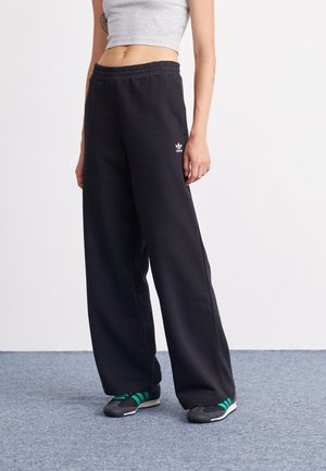 Persoon die zwarte, high-waisted, wijd uitlopende Adidas joggingbroek met elastische tailleband en zwarte sneakers met groene strepen draagt, staande op een grijze vloer.