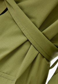 Olivgrön tyg med en mjuk textur, med en inlindad design och synliga stickningsdetaljer. Inkluderar ett knytbälte och en framficka.