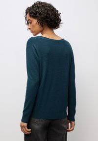 Modrý svetr vyrobený z měkkého materiálu, má široký výstřih, dlouhé rukávy a volnější střih, doplněný o tmavě šedé džíny.