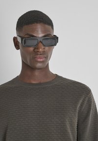 Occhiali da sole neri rettangolari con montatura spessa, indossati sopra un maglione verde scuro e strutturato. Le lenti sono scure e riflettenti.