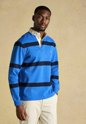 Uomo che indossa una maglia da rugby blu a maniche lunghe con strisce orizzontali nere e pantaloni beige, in piedi su uno sfondo beige uniforme.