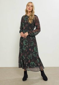 Zwarte maxi-jurk met bloemenpatroon en lange doorzichtige mouwen, met groene en roze bloemenprints. Draagt met zwarte enkellaarsjes. Gladde, lichtgewicht stof.