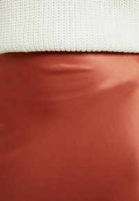 Jupe en satin couleur rouille avec une texture lisse, présentant un design ajusté. Association avec un haut en tricot côtelé crème, partiellement visible en haut.