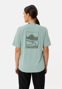 Chemise à manches courtes vert clair en tissu respirant. Présente un design graphique au dos représentant des montagnes et un sentier sinueux.