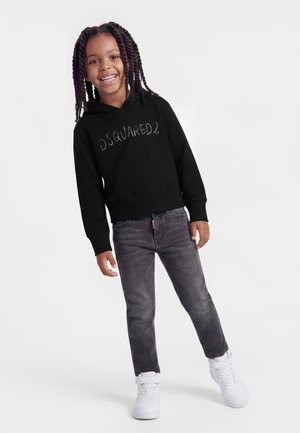 Lächelnes Kind mit geflochtenem Haar, das einen schwarzen DSQUARED2-Hoodie, dunkelgraue Jeans und weiße Turnschuhe trägt, stehend vor weißem Hintergrund.