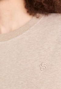 Beige genser med teksturert stoff, rund hals og en liten, brodert logo på brystet.