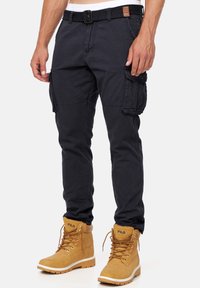 Pantalones cargo en azul marino oscuro con un corte slim, que cuentan con múltiples bolsillos laterales y un cinturón tejido. Combinados con botas beige con cordones.