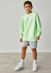 Groene sweatshirt met blauwe tekst, gecombineerd met grijze shorts met een logo. Compleet met witte sneakers en enkelsokken, getoond op een model.