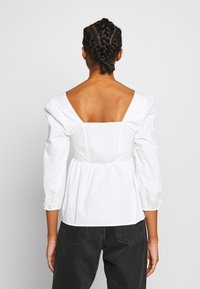 Vit peplumblus med fyrkantig rygg, puffärmar och smockdetaljer vid midjan. Matchad med mörka denimbyxor.