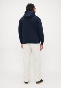 Uomo con capelli intrecciati che indossa un hoodie blu navy, pantaloni avorio e scarpe da ginnastica nere, in piedi di spalle contro uno sfondo semplice.