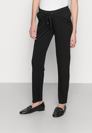 Kobieta w czarnych, dopasowanych spodniach ze ściągaczem i czarnych skórzanych loafersach, stojąca na tle jednolitego jasnego tła.