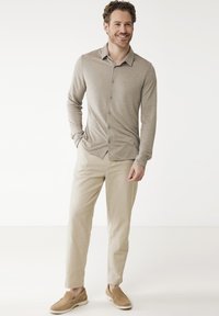 Chemise beige à manches longues, boutonnée, avec une texture douce ; associée à un pantalon tapered beige clair et des chaussures slip-on beiges, sur un fond uni.