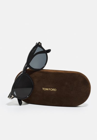 Ochelari de soare negri cu lentile întunecate și rame rotunde, așezați pe o carcasă din suede maro. Logo-ul auriu "TOM FORD" pe carcasa.