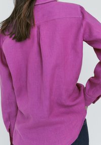 Fuchsia linen shirt with a back yoke and long sleeves, featuring a relaxed fit and subtle texture. Hand on hip, partially tucked into dark jeans.