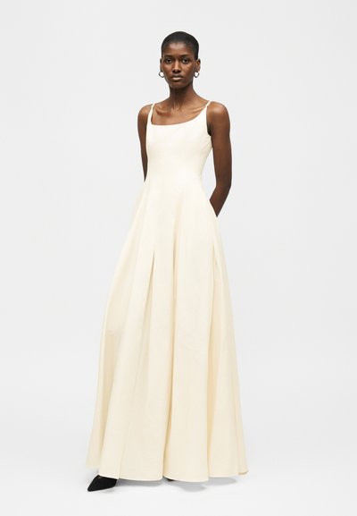 Γυναίκα που φοράει αμάνικο, μακρύ φόρεμα εκρού χρώματος με εφαρμοστό μπούστο και φαρδύ φόρεμα, στέκεται με τα χέρια πίσω από την πλάτη της.