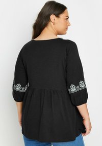 Blusa nera con maniche a sbuffo a tre quarti e ricami floreali bianchi ai polsini. Cucitura arricciata e tessuto morbido e leggero.