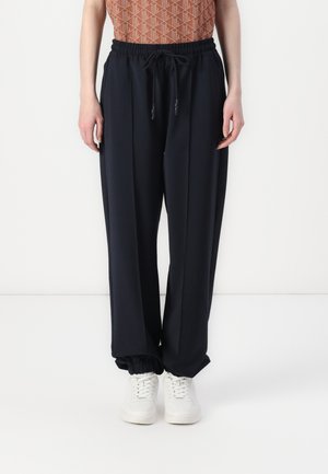 Pantaloni della tuta blu marino con caviglia elastica e vita elastica, dotati di un cordino, abbinati a sneakers bianche su uno sfondo neutro.