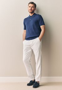 Dunkelblaues Polohemd mit kurzen Ärmeln, weiße Leinenhose und dunkle Schuhe. Der Stoff wirkt weich, mit glatter Textur und einfarbigem Design.