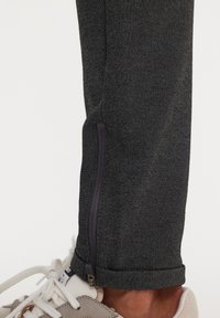 Joggers negros con textura y cremallera en el tobillo, combinados con zapatillas grises y blancas. La vista en primer plano resalta los detalles de la tela y el cierre.