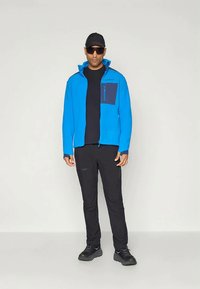 Niebieska polarowa kurtka z granatową kieszenią, czarna koszulka, czarne spodnie i czarne buty. Model ma na sobie okulary przeciwsłoneczne i czapkę w kolorze czarnym.