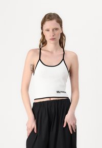 Hvit ribbet crop topp med svart kant og logodetalj nederst. Tynne stropper og tettsittende design. Modellen har på seg svarte joggebukser med snøring.