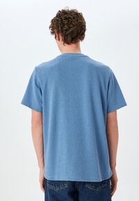 Blå tekstureret kortærmet t-shirt med rund halsudskæring, der viser bagsiden og en let buet kant. Kombineret med blå denimjeans.