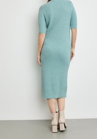 Femme portant une robe midi en maille côtelée couleur sarcelle avec des manches jusqu'au coude et des bottines beiges à talons carrés, debout contre un mur blanc.