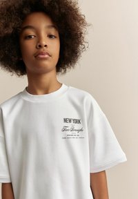 Hvid t-shirt med korte ærmer, der har et grafisk print med teksten "NEW YORK" og "Five Boroughs" i sort. Blødt bomuldsmateriale.