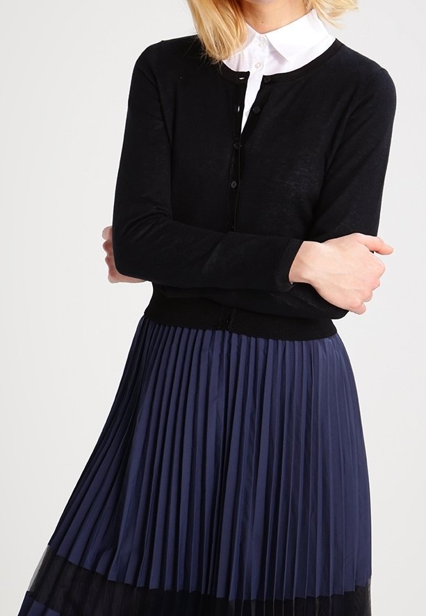 Femme portant un cardigan noir boutonné sur une chemise blanche à col et une jupe plissée bleu marine avec des détails noirs.