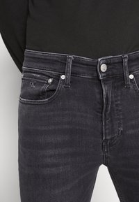 Primer plano de unos vaqueros de mezclilla gris oscuro con el logo de Calvin Klein en el botón y un pequeño bolsillo frontal, llevados con una parte superior negra.