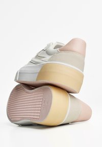 Sneakers bianchi con un tallone rosa pallido e beige e suola testurizzata, impilati per mostrare il profilo posteriore e i dettagli del design della scarpa.