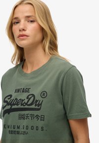 Grünes Baumwoll-T-Shirt mit Rundhalsausschnitt und kurzen Ärmeln, mit schwarzer Textdruckschrift "Vintage SuperDry" auf der Vorderseite.