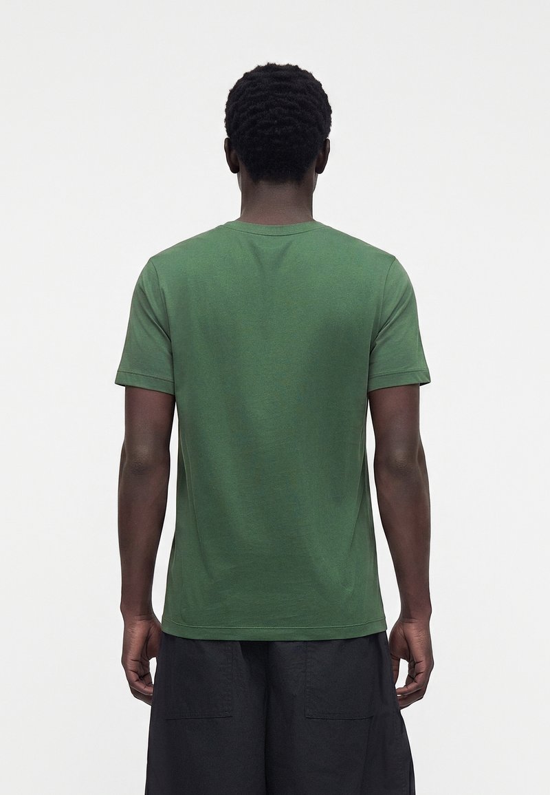 Mand med kort krøllet hår iført en ensfarvet grøn kortærmet T-shirt og sorte shorts, vendt væk fra kameraet mod en hvid baggrund.