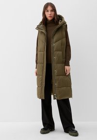 Olijfgroene gewatteerde vest met een capuchon, voorzien van een ritssluiting, voorknopjes en zijzakken. Gedragen over een bruine coltrui en zwarte broek.
