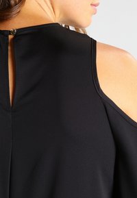 Svart topp med kallaxel-design, slät tyg och en nyckelhålsknäppning med en liten accentknapp.