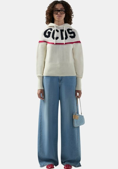 Donna con capelli ricci e occhiali che indossa un maglione bianco GCDS, jeans blu a gamba larga, scarpe rosse e tiene in mano una piccola borsa blu.
