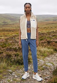 Mujer de pie en un sendero rocoso con chaleco beige de forro polar, top deportivo estampado, pantalones jogger azules y zapatillas blancas en un paisaje exterior nublado.