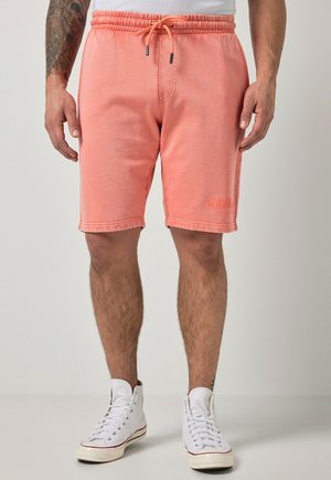 Mand iført koralfarvede snørrebukser og hvide høje sneakers står mod en ensfarvet baggrund.