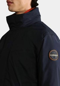 Giacca invernale blu navy con colletto alto, texture liscia e una patch rotonda con logo sul braccio sinistro. Tasca anteriore visibile.