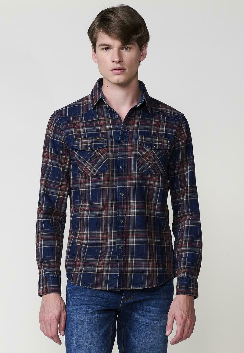 Camicia a maniche lunghe a quadri in blu navy, rosso e crema. Presenta due tasche sul petto, colletto con bottoni e orlo curvo. Tessuto morbido.